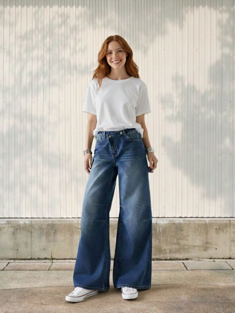 Femme rousse portant un t-shirt blanc et un jean large délavé avec un bouton asymétrique sur fond clair