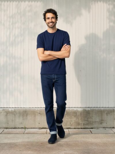 Homme type caucasien brun portant un t-shirt bleu marine et bottine assortie avec un jean droit bleu brut 100% coton avec un petit bord relevé sur fond éclairé.