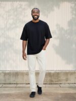 Un homme de couleur portant un t-shirt noir oversize et un jean droit blanc au style minimal et épuré dans un lieu ensoleillé.
