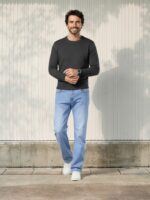 Homme caucasien brun se tenant debout dans un lieu très éclairé et portant un pull léger manche longs gris et un jean droit bleu clair en denim épais et stretch.