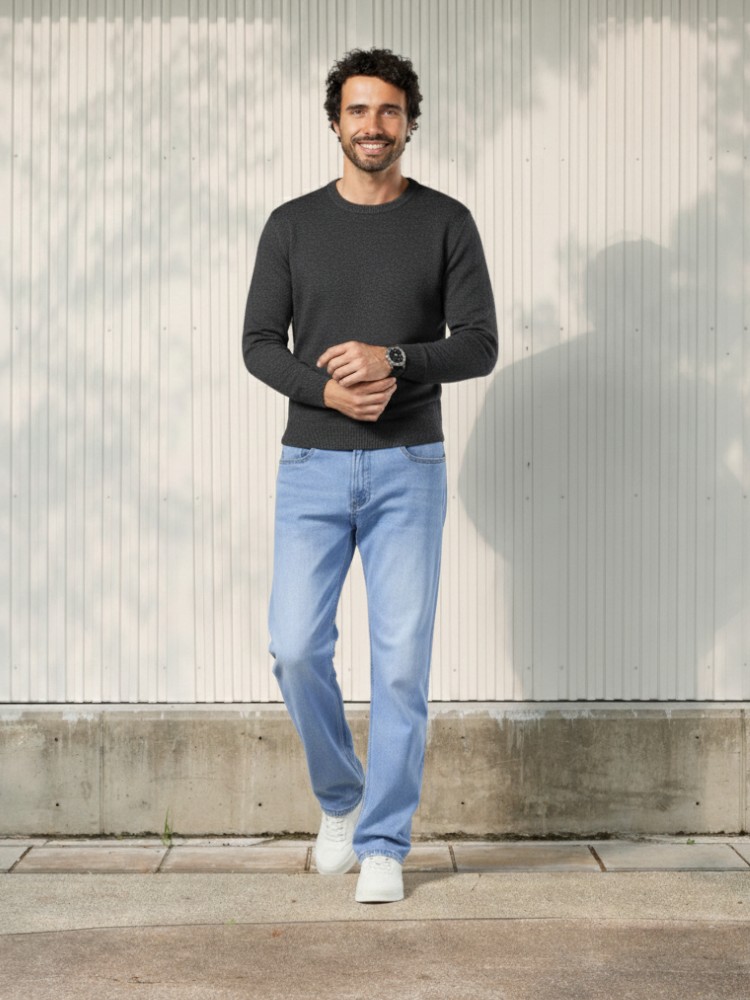Homme caucasien brun se tenant debout dans un lieu très éclairé et portant un pull léger manche longs gris et un jean droit bleu clair en denim épais et stretch.