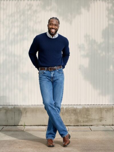 Homme de couleur, les mains dans le dos et jambe croisées se tenant debout, vêtu d'un pull léger bleu marine, d'une ceinture marron assortie aux chaussures et d'un jean droit bleu en denim stretch élégant sur fond clair et ensoleillé.