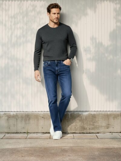 Homme caucasien chatain clair adossé contre un mur dans un lieu tres eclairé portant un pull leger gris et un jean droit bleu foncé denim épais et des sneakers blanches.