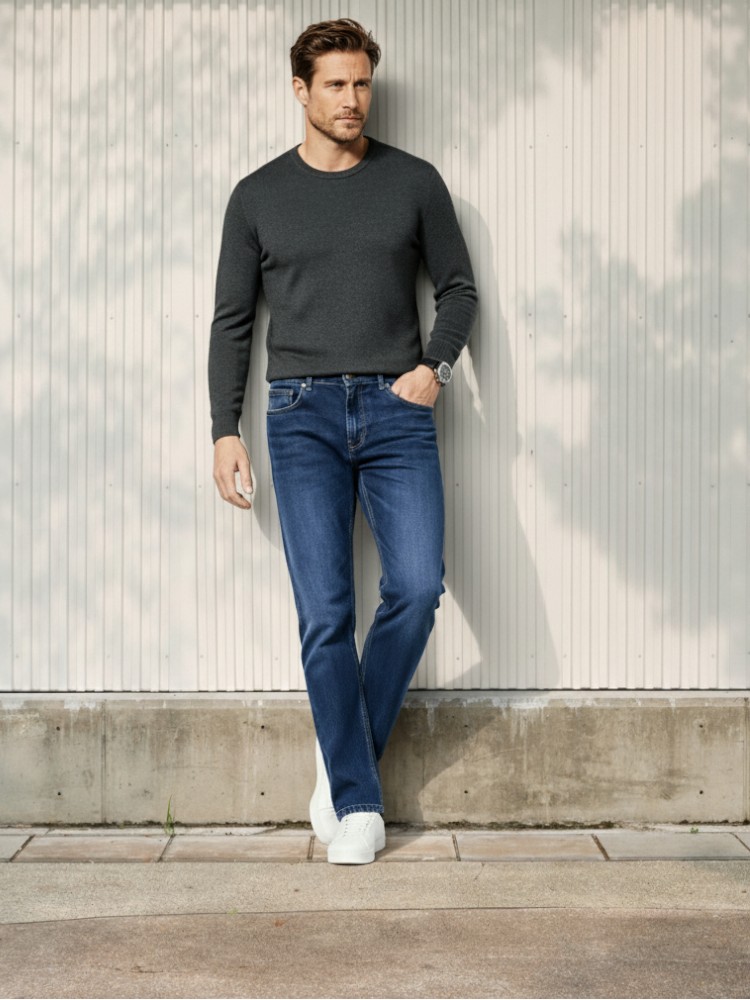 Homme caucasien chatain clair adossé contre un mur dans un lieu tres eclairé portant un pull leger gris et un jean droit bleu foncé denim épais et des sneakers blanches.