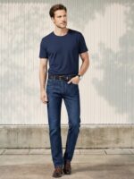 Homme caucasien plain-pied avec une main dans la poche, le visage tourné sur le gauche, habillé d'un t-shirt bleu marine et d'un jean droit bleu marine en coton stretch dans un lieu éclairé et ensoleillé.