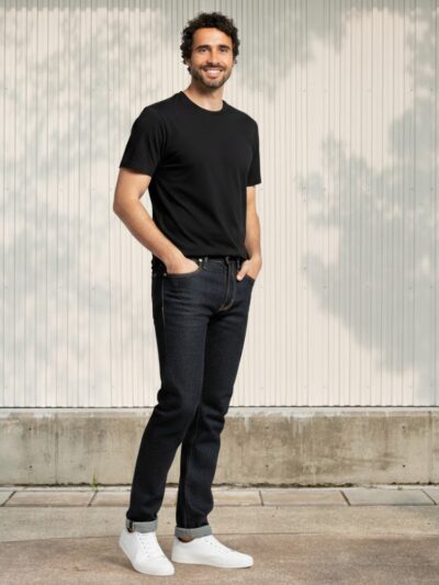 Homme de type caucasien vêtu d'un t-shirt noir et d'un jean droit brut dark blue épais avec des sneakers blanches.