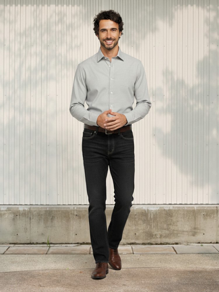 Homme caucasien brun portant une chemise manche longue grise et un jean droit nor stretch élégant et souple avec chaussure marron dans un lieu ensoleillé.