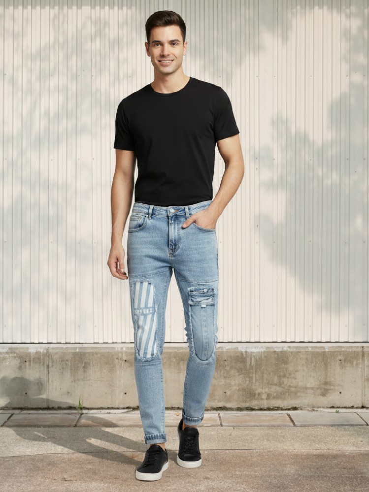 Jeune homme caucasien brun portant un t-shirt noir et un jean fuselé bleu clair délavé avec patchwork dans un lieu éclairé et ensoleillé.