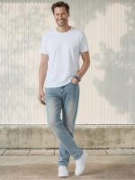 Homme caucasien avec un t-shirt blanc, une main dans la poche, les jambes croisées, habillé d'un jean regular bleu clair délavé esprit rétro sur fond clair ensoleillé.
