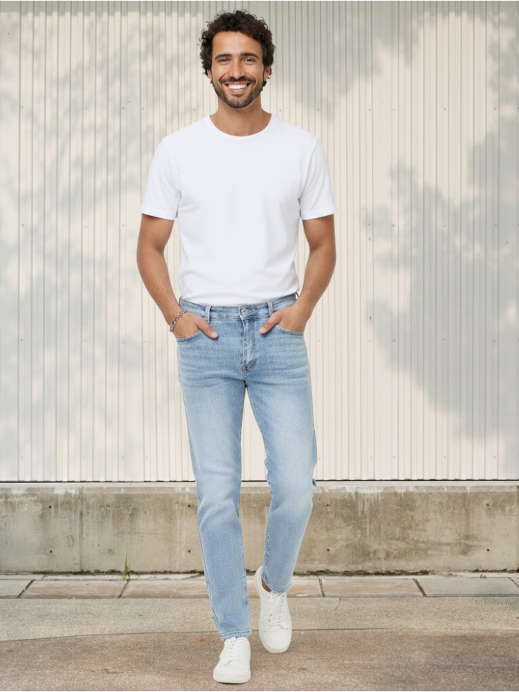 Homme caucasien châtain avec un t-shirt blanc et un jean regular bleu clair effet délavé en coton stretch sur fond clair et ensoleillé.