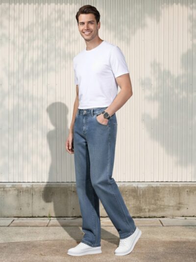 Homme caucasien légèrement de profil vêtu d'un t-shirt blanc, d'une paire de sneakers blanche et d'un jean regular bleu, coupe droite au style vintage dans un lieu ensoleillé.