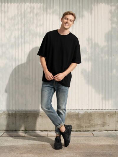 Jeune homme blond avec t-shirt oversize noir et jean regular bleu patiné revers contrastants dans un lieu éclairé et ensoleillé.