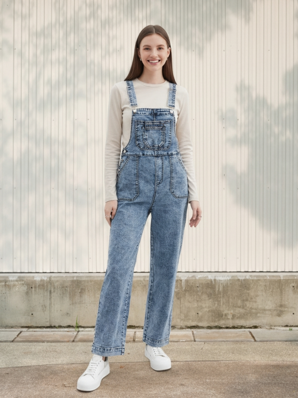 Femme portant un haut blanc à manches longues avec une salopette en jean bleu à coupe droite, sur fond clair