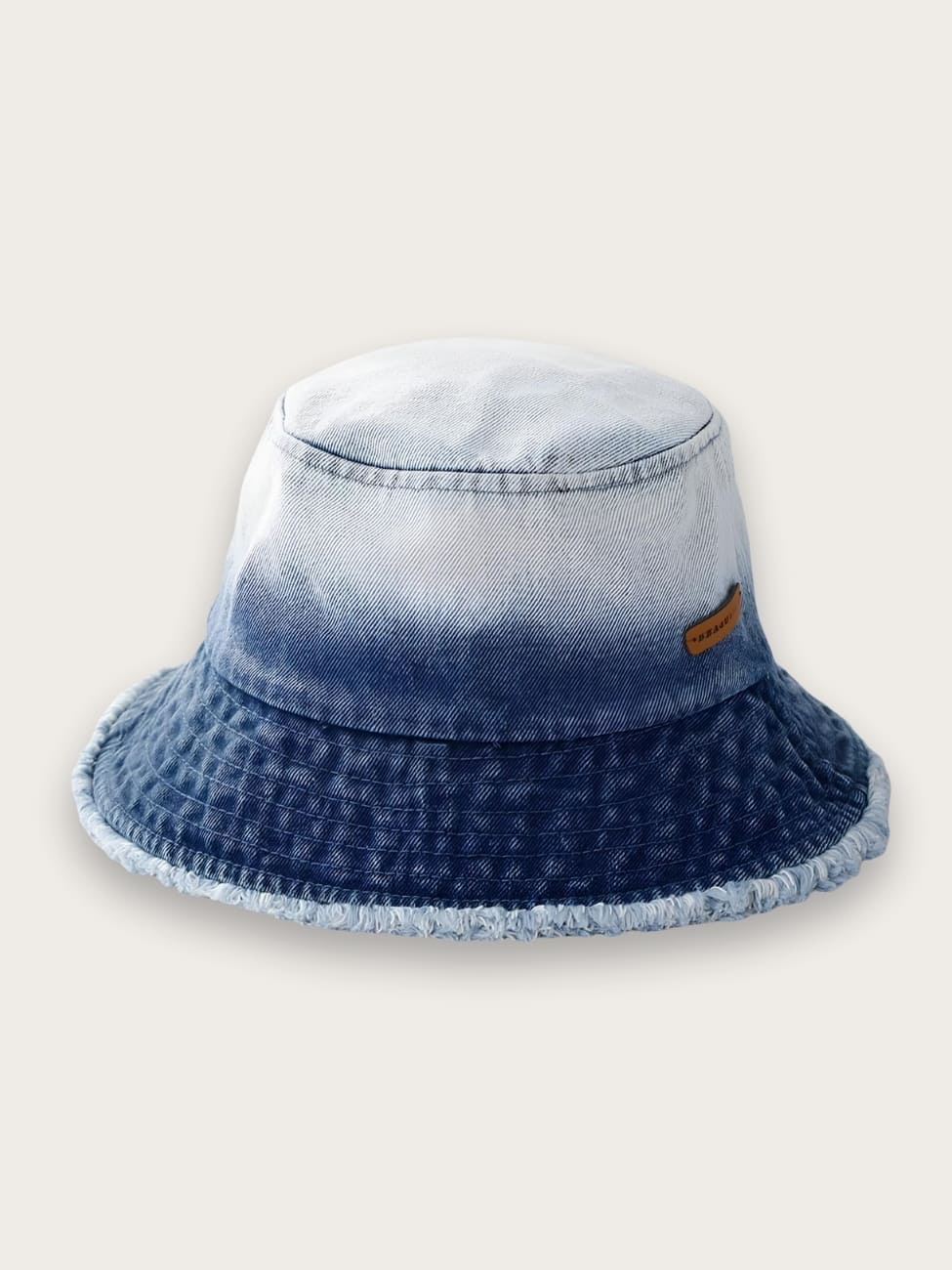 Bob en jean effet délavé et effiloché pour femme. La couleur est dégradé du bord extérieur au haut du bob, du bleu foncé au bleu très clair. Le bord est légèrement effiloché. Sur fond beige.