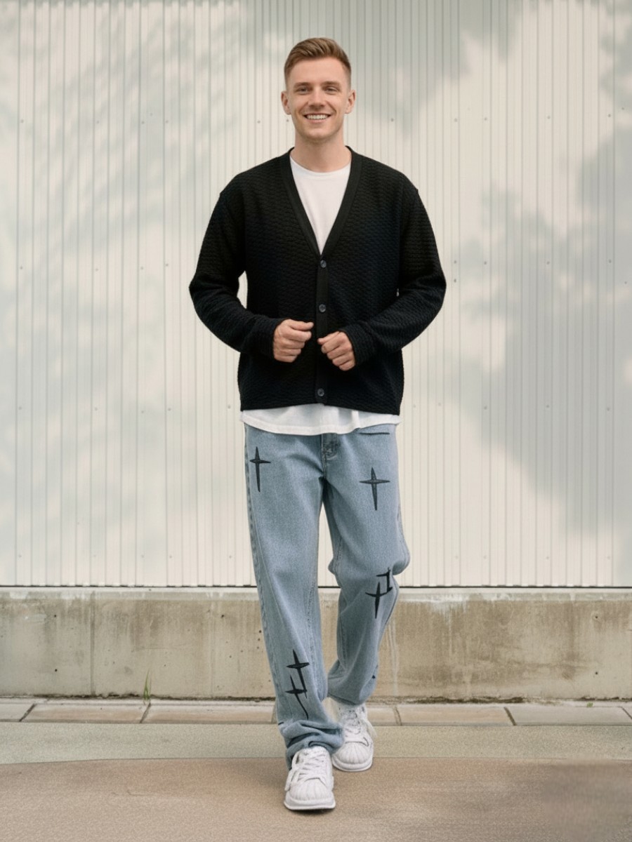 Un jeune homme habillé d'un gilet noir, d'un t-shirt blanc, et d'un jean baggy bleu clair aux détails brodés dans un lieu éclairé et ensoleillé.