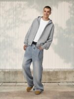 Jeune homme vêtu d'un jean baggy gris clair délavé, d'un t-shirt blanc et d'un gilet à capuche gris sur fond ensoleillé.