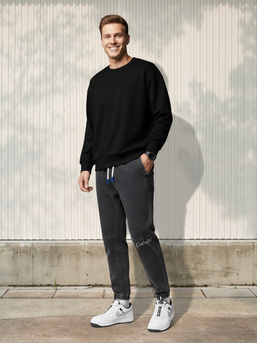 Homme caucasien portant un pull noir, des sneakers blanches avec motifs noirs et un jean élastique taille et cheville à la coupe jogger, sur fond clair et ensoleillé.