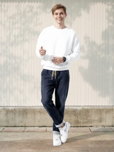 Homme caucasien en plain-pied, vêtu de sneakers blanches, d'un pull blanc et d'un jean taille élastique en denim brut, coupe fuselée avec cordons réglables, sur fond clair et ensoleillé.