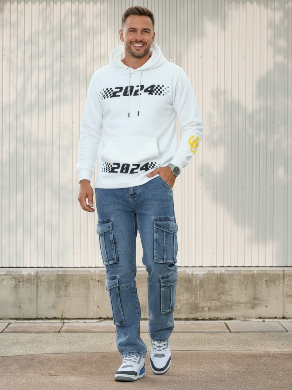 Un homme souriant vêtu d'un sweat avec imprimés, d'un jean cargo bleu multi-poches dans un lieu clair et ensoleillé.