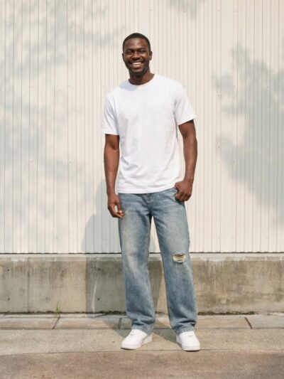 Homme portant un t-shirt blanc avec un jean bleu délavé déchiré, à coupe droite, sur fond beige