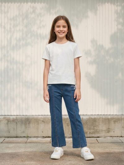 Petite fille portant un t-shirt blanc avec un jean bleu foncé évasé avec des broderies, sur fond clair