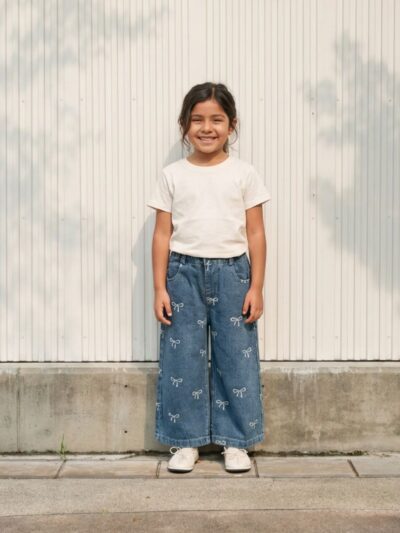 Petite fille portant un t-shirt blanc avec un jean bleu à broderie noeuds, sur fond clair