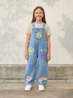 Petite fille portant une salopette en jean bleu avec des motifs fleurs, coeurs et arc-en-ciel, sur fond clair