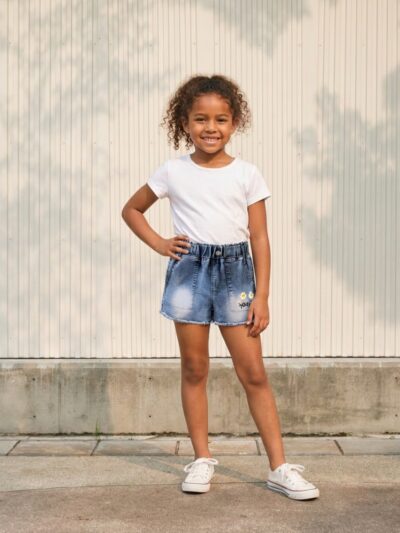 Petite fille portant un t-shirt blanc avec un short en jean bleu à broderies marguerite, sur fond clair