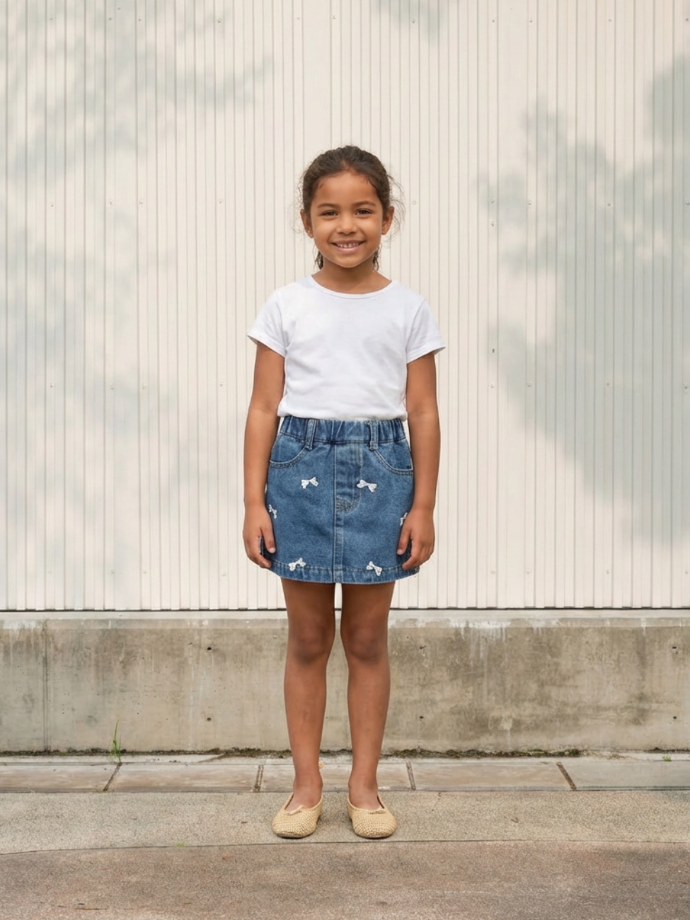 Petite fille portant un t-shirt blanc avec un short en jean bleu à broderie noeuds, sur fond beige