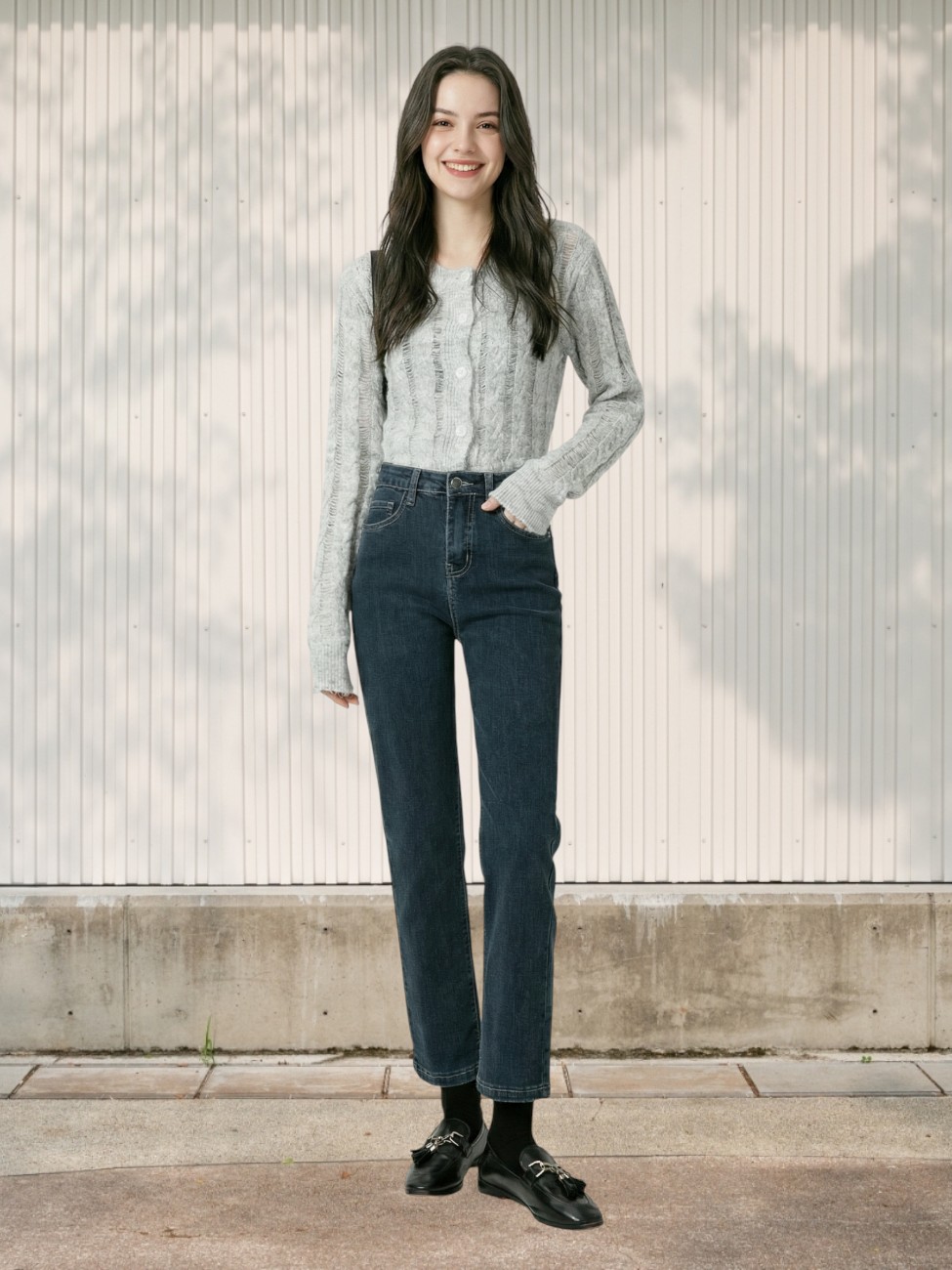 Une jeune femme souriante portant un Jean 7/8 femme taille haute coupe droite en denim bleu foncé avec un gilet gris clair, posant devant un mur blanc strié où se reflètent des ombres de feuilles d'arbres.