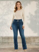 Femme portant un haut blanc avec un jean push up bleu foncé délavé à coupe bootcut, sur fond beige