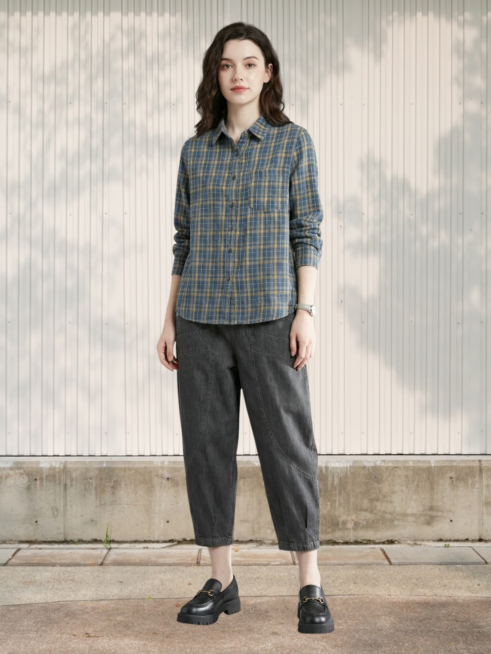 Femme debout portant le Jean 7/8 femme design ample à poches marquées : coupe large, tissu gris foncé, poches visibles, chemise à carreaux et mocassins noirs.