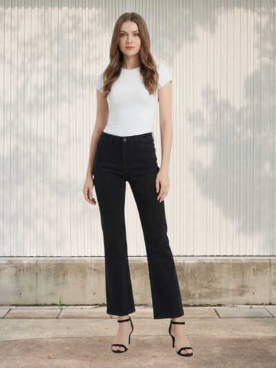 Femme debout portant un Jean noir 7/8 coupe droite : taille haute, jambes légèrement évasées, silhouette élancée, look chic et minimaliste avec talons fins.
