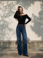 Femme souriante debout en extérieur, portant un Jean femme taille basse flare bleu délavé wide avec un haut noir épaules nues, jambes allongées par la coupe flare.