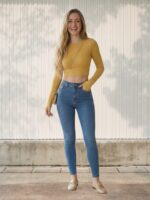 Femme debout souriante portant un Jean taille haute push-up effet sculptant bleu clair avec un top court jaune moutarde et un sac en bandoulière, posant devant un mur blanc strié.