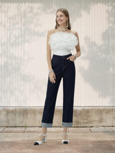 Jeune femme souriante en Jean femme taille haute droit denim foncé avec revers, assorti d’un bustier blanc à volants, posant devant un mur clair.